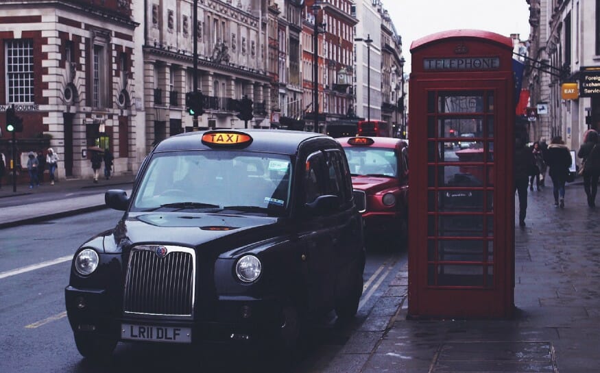 A red and black London taxi cab parked next to a telephone booth